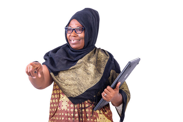 Muslim mature businesswoman wearing glasses, holding laptop and pointing forward, smiling.