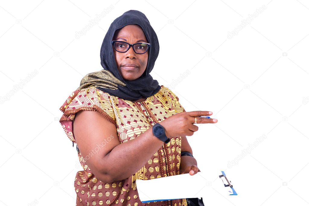 Muslim mature woman wearing glasses, holding clipboard and pointing finger aside while looking at camera.