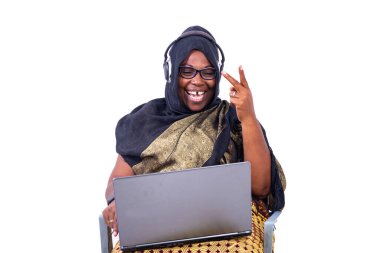 Güzel, gülen Müslüman iş kadını beyaz arka planda oturuyor. Laptop ve kulaklık kullanarak iletişim kuruyor. Zafer jestleri yapıyor..