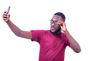 Gözlüklü genç bir adam beyaz arka planda durup gülümserken cep telefonuyla kendi fotoğrafını çekiyor..