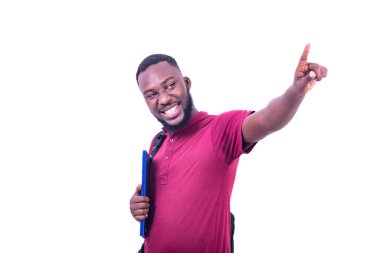 Beyaz bir arka planda duran tişörtlü genç bir öğrenci sırt çantası giyiyor ve kitapları parmağıyla gülümsüyor..