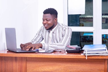 Çizgili gömlekli genç bir işadamı ofiste oturmuş dizüstü bilgisayarda gülümsüyor.