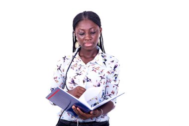 Saçları örgülü genç bir kadın beyaz bir arkaplanda durup defterini kontrol ediyor..