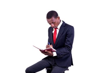 Ceketli bir işadamı beyaz arka planda bir taburede oturuyor ve bir günlüğe yazıyor..