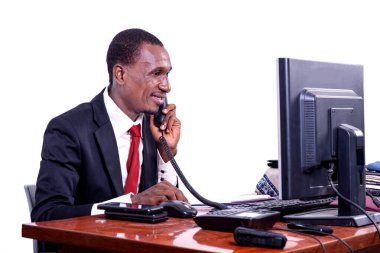 Yakışıklı, yetişkin iş adamı ofiste bilgisayar karşısında oturuyor ve gülümserken cep telefonuyla konuşuyor..