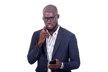 Beyaz arka planda duran ceketli genç bir işadamı cep telefonuyla düşünürken.