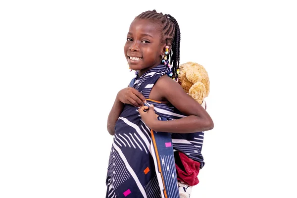かわいい女の子は伝統的なアフリカのふんどしを着てカメラで微笑みながら背中にテディベアを担いで — ストック写真