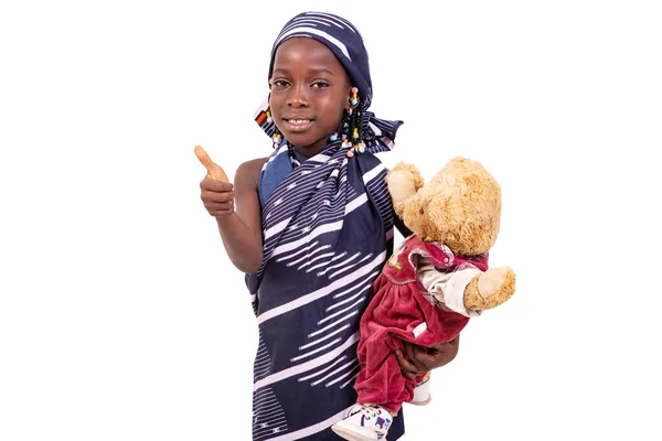 かわいい女の子は伝統的なアフリカのふんどしを着てテディベアを持って親指を立てて笑っています — ストック写真