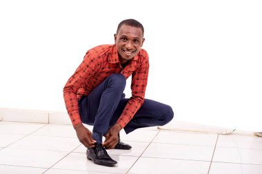 Kareli gömlekli yakışıklı bir işadamı beyaz bir arka planda çömelmiş, bağcıklarını düzeltiyor, gülümsüyor..