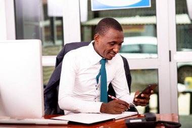 Genç bir işadamı ofisinde oturmuş telefonda bir mesaj okuyor ve gülümsüyor..