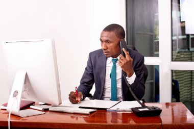 İşadamı bilgisayarın önünde oturuyor ve ofiste notlar alırken cep telefonuyla konuşuyor..