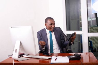 Takım elbiseli bir işadamı masada oturmuş tablete bakıyor ve zafer jesti yapıyor..