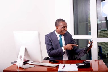 Takım elbiseli bir işadamı masada oturmuş tableti işaret ediyor ve gülüyor..