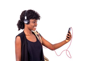 Beyaz arka planda duran sırt çantalı genç bir öğrenci cep telefonu ve kulaklık kullanarak müzik dinliyor.