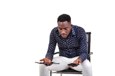 Genç sakallı bir adam beyaz arka planda oturuyor elinde tablet ve cep telefonu var.