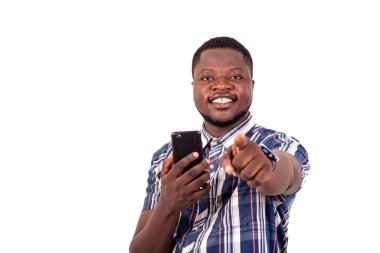 Kısa kollu tişörtlü genç bir adam beyaz arka planda cep telefonuyla gülümsüyor..