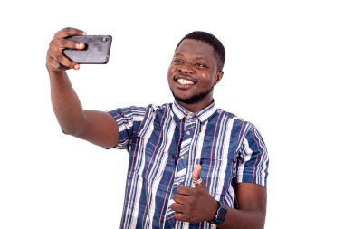 Kısa kollu gömlekli genç bir adam beyaz arka planda duruyor ve cep telefonuyla video görüşmesi yapıyor ve gülümsüyor..