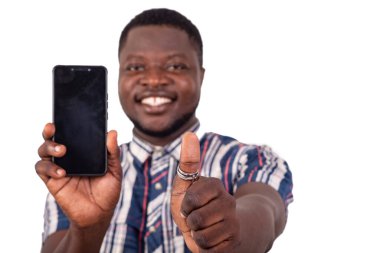 Kısa kollu gömlekli genç bir adam beyaz arka planda duruyor cep telefonunu sunuyor ve gülümserken iyi bir hareket gösteriyor..