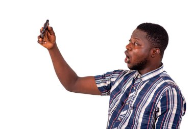 Kısa kollu gömlekli genç bir adam beyaz arka planda duruyor ve cep telefonuyla video görüşmesi yapıyor..