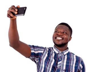 Kısa kollu gömlekli genç bir adam beyaz bir arka planda durup cep telefonuyla fotoğraf çekiyor, gülümsüyor..