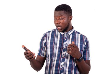 Beyaz arka plan üzerinde duran gömlekli genç bir adam şaşırmış bir şekilde cep telefonuna bakıyor..