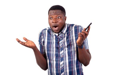 Beyaz arka plan üzerinde duran tişörtlü genç bir adam şok olmuş bir şekilde elinde cep telefonu tutuyor..