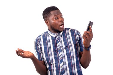 Beyaz arka planda duran gömlekli genç bir adam şok olmuş bir şekilde cep telefonuna bakıyor..