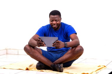 Spor kıyafetleri giymiş yakışıklı bir gencin portresi yere oturmuş, gülümseyen dijital bir tablet kullanıyor..