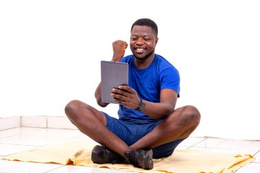 Spor kıyafetleri giymiş yakışıklı bir gencin portresi yere oturmuş dijital tablet tutarken kolları havada kazanan bir jest yapıyor..