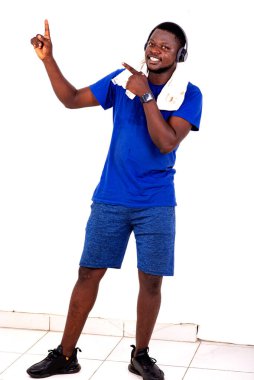 Spor kıyafetli genç adam, kulaklıkları ve havlusuyla spor salonunda durup parmaklarını göstererek kameraya bakıp gülümsüyor..