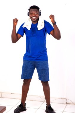 Boynunda havluyla salonda dikilmiş, müzik dinlerken zafer jestleri yapan genç bir adam spor üstüne kulaklık takıyor.