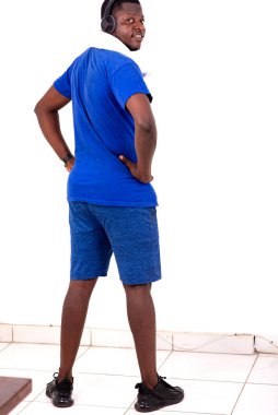 Spor kıyafetli genç adam, beyaz arka planda, kulaklıklarla kameraya gülümseyerek bakıyor..