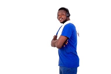 Spor giyimli, kollarını çapraz bağlamış kulaklıklı, beyaz arka planda duran ve kameraya bakıp gülümseyen genç bir adam..