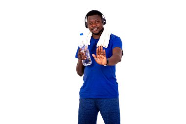 Boynunda havluyla, kulaklık takan, plastik su şişesi tutan ve gülümserken avuç içi gösteren genç sporcu..