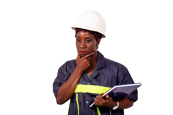 仕事用の制服を着た美しい女性技術者の肖像ノートを持ち顎の下に手を挙げて考える白いハード帽子 — ストック写真