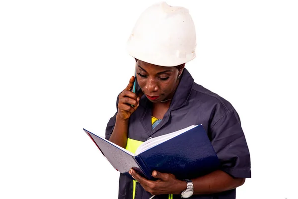 携帯電話で話す仕事の制服と白い硬い帽子を身に着けている美しい女性エンジニアの肖像画やノートブックでノートをチェック — ストック写真
