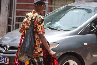 Abidjan, Fildişi Sahili - 10 Nisan 2025: Siyah şapkalı bir sokak satıcısı, omzunda siyah plastik bir çanta, sokakta bağırarak yürüyor.