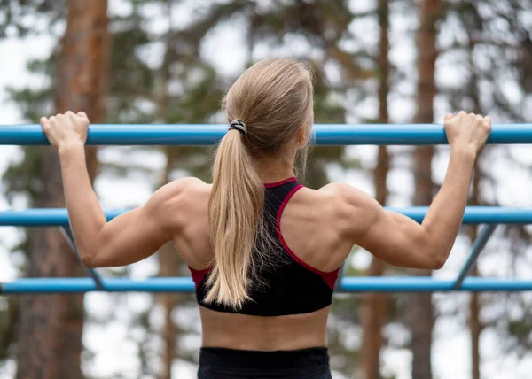 Atletyczna blondynka piękna biała kobieta robi podbródek ups ćwiczenia na boisku sportowym zewnątrz w lecie, górnej części ciała, zbliżenie, selektywne skupienie — Zdjęcie stockowe