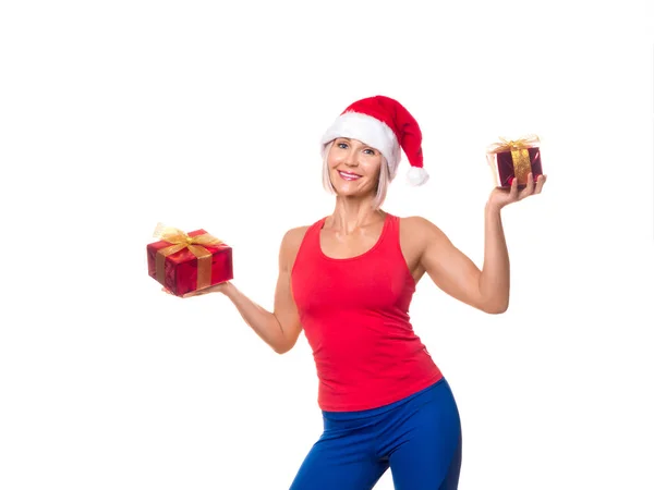 Świąteczna wyprzedaż lub prezent w klubie fitness. Uśmiechnięta, wysportowana, dorosła kobieta w czerwonej odzieży sportowej i czapce Mikołaja z pudełkami z prezentami w dłoniach robi promo, izolowane na białym. — Zdjęcie stockowe