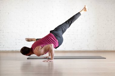 Male yoga practice, advanced level. Caucasian fit man doing side handstand with one leg up in fitness loft studio indoor, selective focus.