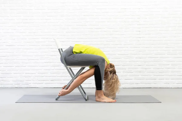 Sandalyeli yoga. Kafkasyalı kadın oturur ve arkada çalışır ve kalçaları paspaslarla sallanır. Stüdyoda, seçici bir odak noktası vardır..