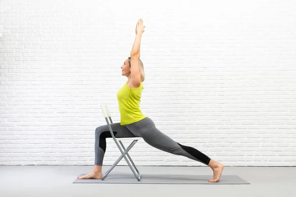 Sandalyeli yoga. Kafkasyalı kadın, beyaz çatı katındaki stüdyo dairede sahne malzemesi kullanarak savaşçı pozu veriyor..