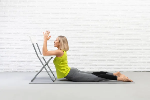Yoga içeride, sahne donanımlı, geriye doğru geriniyor. Çekici beyaz kadın yerde yatar ve dirseklerini bir sandalyeye koyar..