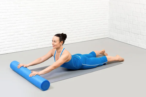 Back extention drill with foam roller. Adult smiling caucasian woman in blue sportswear lies on her stomach and does pilates workout in loft fitness studio, indoor. — Stock Photo, Image