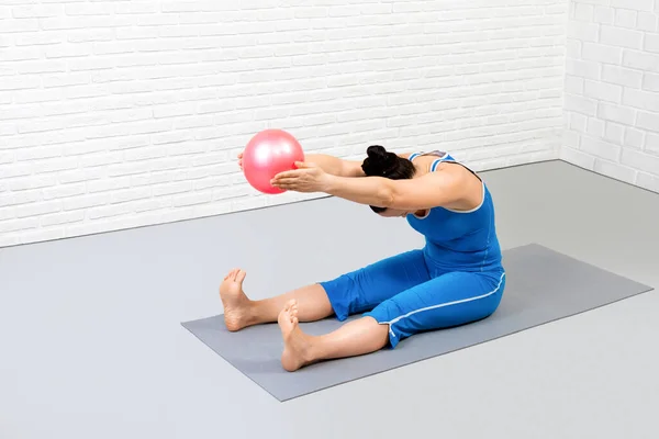 Gerinc nyújtás előre fúró kis illeszkedő labdát. Felnőtt kaukázusi nő kék sportruházatban pilates tetőtéri fitness stúdió beltéri. — Stock Fotó
