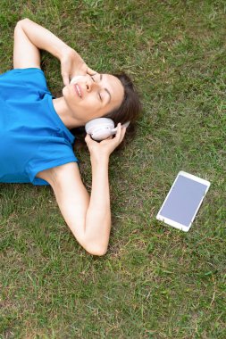 Teknolojiyle rahatla. Büyük kulaklıklı, mutlu, beyaz bir kadın yazın açık hava parkında tabletinin yanındaki çimlerin üzerine uzanmış, dikey fotoğraf, seçilmiş odak.