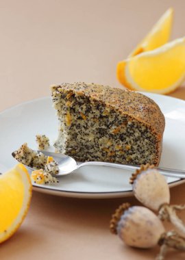 Haşhaş tohumu ve portakal kabuğu vejetaryen yumurtasız ve sütsüz kek, yanında mavi keten tekstil ve haşhaş kafası olan turuncu tabağı ısır, yakın plan, kopyalama alanı, vejetaryen yemeği ve hamur işi kavramı