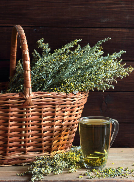 Wormwood blooming herb, absinth plant in a wicker on wooden rustic background with cup of herbal tea, closeup, copy space, alternative medicine naturopathy and alcohol production concept