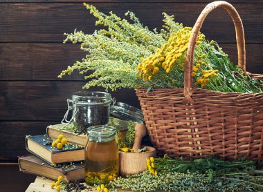 Herbal helminthic triad: absinth, tansy and cloves; fresh herbs in the wicker basket and wooden mortar, on old books,  healing infusion is nearby, closeup, copy space, antihelminthic medicine concept clipart