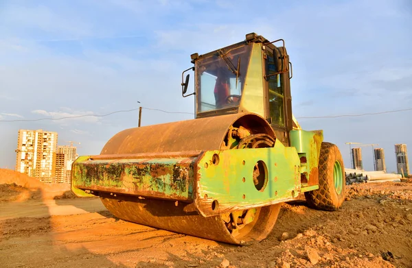 Vibro Roller Soil Compactor leveling ground at construction site. Vibration single-cylinder road roller on construction road. Road work for new asphalt laying. Tower cranes build high-rise buildings